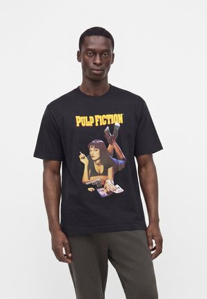 Homme portant un t-shirt noir avec texte jaune "Pulp Fiction" et un dessin d'une femme allongée tenant une cigarette, un pistolet et des magazines.