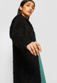 Cardigan noir en peluche avec des manches longues, ouverture devant et surface texturée. Le mannequin porte une robe verte en dessous.