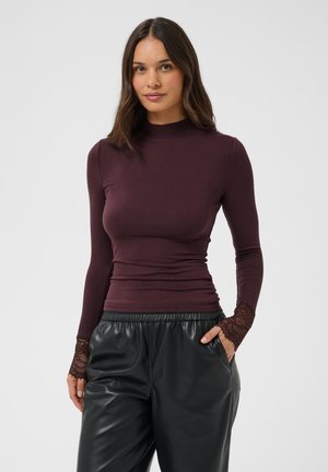 Femme aux cheveux foncés portant un top bordeaux cintré à manches longues avec des poignets en dentelle et un pantalon en cuir noir ample, mains dans les poches.