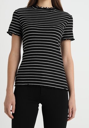 Mujer con una blusa negra de manga corta con rayas horizontales blancas y bordes con volantes, combinada con pantalones negros, sobre un fondo liso.