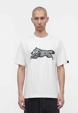 Homme à tête rasée portant un t-shirt blanc avec un graphisme animal stylisé noir, debout devant un fond uni.