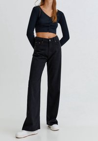 Jeans de pierna ancha negros con remaches de cobre y cintura alta, combinados con una blusa negra ajustada de manga larga con cierre de botones. Zapatillas blancas.