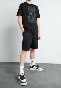 Camiseta negra con un diseño gráfico en verde y gris, combinada con pantalones cortos de malla negros y zapatillas negras y blancas con detalles.