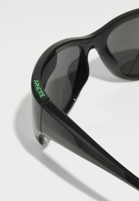 Black sunglasses with sleek plastic frames, dark lenses, and a green "ARNETTE" logo on the side. Smooth texture and modern design.