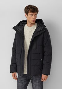 Schwarze Steppjacke mit Kapuze, ausgestattet mit einem Frontreißverschluss, großen Taschen und einem gesteppte Design. Darüber getragen wird ein heller, cremefarbener Pullover mit Struktur.