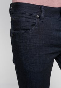 Jeans de mezclilla azul oscuro con un diseño clásico, costuras resistentes, bolsillos funcionales y una textura suave.