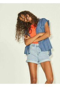 Džínová košile s knoflíky a ohrnutými rukávy, kombinovaná s jasně oranžovým topem a světle modrými džínovými šortkami, které mají manžety a volný střih.