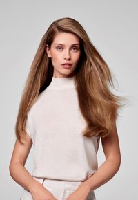 Modella con lunghi capelli castano chiaro, lisci, che indossa un top beige chiaro a maniche corte e collo alto. La pelle è chiara e l'espressione è neutra.