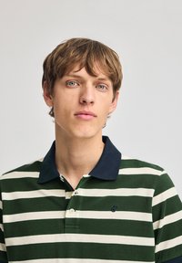 Grön- och vitrandig polo skjorta med en marinblå krage, korta ärmar och en liten logotyp på bröstet. Tyget verkar ha en texturerad yta.