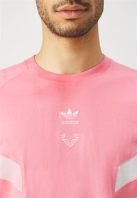 Roza pamučna majica s okruglim izrezom, koja sadrži bijeli vezeni Adidas logotip i grafički dizajn u središtu. Kratki rukavi s detaljima naglaska.