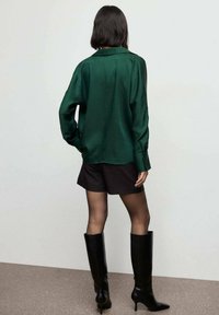 Camisa larga de satén verde con mangas largas y puños abotonados, llevada sobre pantalones cortos negros. Botas altas de charol negras, con textura suave y diseño minimalista.