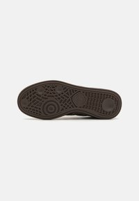 Semelle de sneaker marron en caoutchouc texturé, présentant des motifs complexes et des designs circulaires. Logo clairement visible au centre.