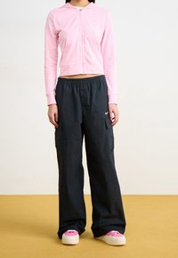 Person som bär en rosa cropped zip-up hoodie, lösa svarta cargobyxor och rosa och vita platåskor som står på ett gult golv.