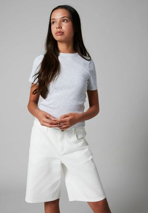 Junge Frau mit langen dunklen Haaren, die ein hellgraues kurzärmeliges Shirt und weiße kniehohe Shorts trägt, stehend mit zusammengelegten Händen.