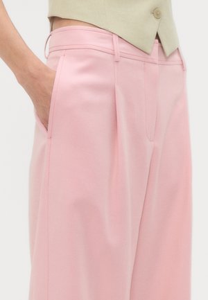 Nahaufnahme einer Person, die hoch taillierte, weit geschnittene rosa Hosen trägt, mit einer Hand in der Seitentasche und einem beigefarbenen, kurz geschnittenen Oberteil.