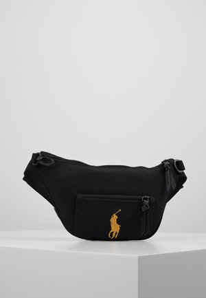 Sac banane noir avec poche zippée à l'avant présentant un logo joueur de polo jaune, sangle réglable et fermeture zippée supérieure sur une surface blanche.
