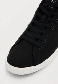 Baskets noires en toile avec des lacets noirs et semelle en caoutchouc blanc à nervures, vue rapprochée des parties avant et latérale.