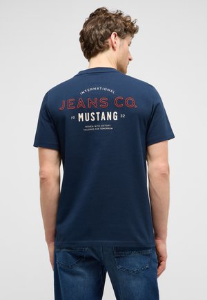 Ein Mann trägt ein dunkelblaues T-Shirt mit dem Schriftzug „Jeans Co. Mustang“ auf dem Rücken und blaue Jeans und steht vor einem schlichten hellgrauen Hintergrund.