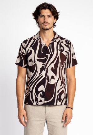 Mężczyzna w koszuli z krótkim rękawem z abstrakcyjnym czarno-beżowym spiralnym wzorem, w połączeniu z jasnobeżowymi spodniami, stoi na białym tle.