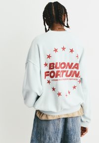 Vaaleansininen ylisuurikokoinen huppari, jossa on punaisia tähtiä ja selässä teksti "BUONA FORTUNA". Hupparissa on rento istuvuus ja resorit hihansuissa.