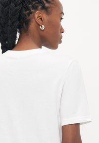 Hvid bomulds t-shirt med korte ærmer, der har en rund halsudskæring og en glat tekstur. Sølv hoops øreringe synlige på modelens øre.