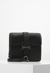 Černá kožená kabelka přes rameno s klopou, detaily s kovovou sponou a řetízkovým popruhem odpočívající na bílé ploše.