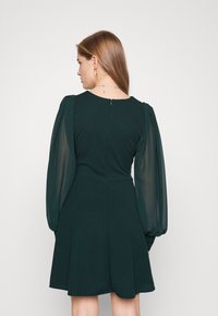 Tummanvihreä mekko, jossa on muotoiltu yläosa ja levenevä helma. Läpinäkyvät puffihihat ja piilotettu takakuvitus. Sulava kankaan pinta.
