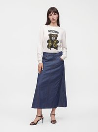 Femme portant un pull blanc Moschino avec un motif ours et une longue jupe en denim bleu, debout avec une main dans sa poche et des talons noirs.