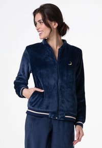 Giacca in pile blu scuro con chiusura a zip, polsini a costine e un accento a forma di luna crescente dorata. Presenta tasche laterali e una texture morbida.