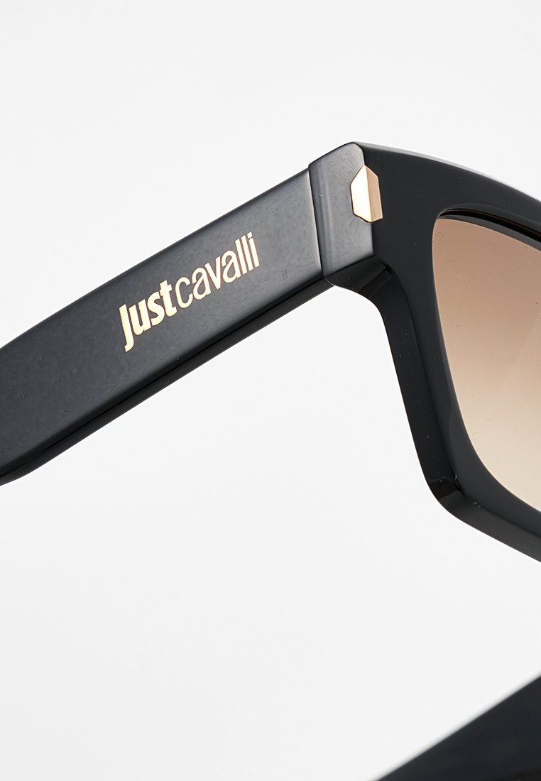 Occhiali da sole neri Just Cavalli con logo dorato sulla tempia e lenti marroni sfumate, su uno sfondo bianco.
