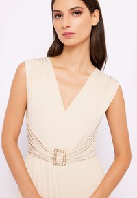 Abito beige con scollatura a V profonda e design senza maniche. Presenta un tessuto arricciato in vita, accentuato da una fibbia dorata e perle.
