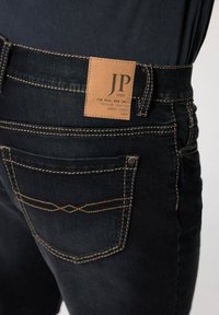 Los jeans de denim oscuro cuentan con una etiqueta de cuero con "JP 1880" en la parte posterior. La costura en contraste resalta el diseño de los bolsillos.