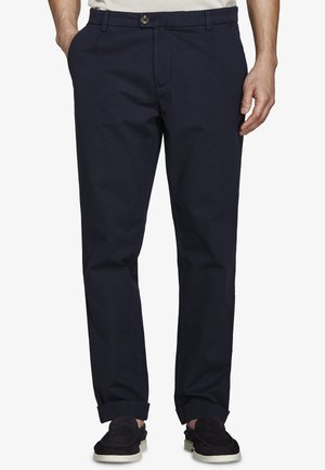 Mężczyzna noszący granatowe chinosy z podwiniętymi nogawkami i czarne wsuwane buty, stojący z jedną ręką w kieszeni na białym tle.