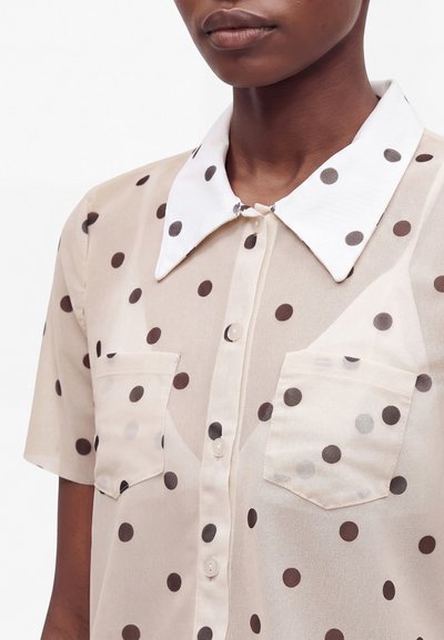 Gros plan d'une personne portant une chemise beige translucide à pois avec un col blanc et des poches avant, montrant les boutons et la texture du tissu.