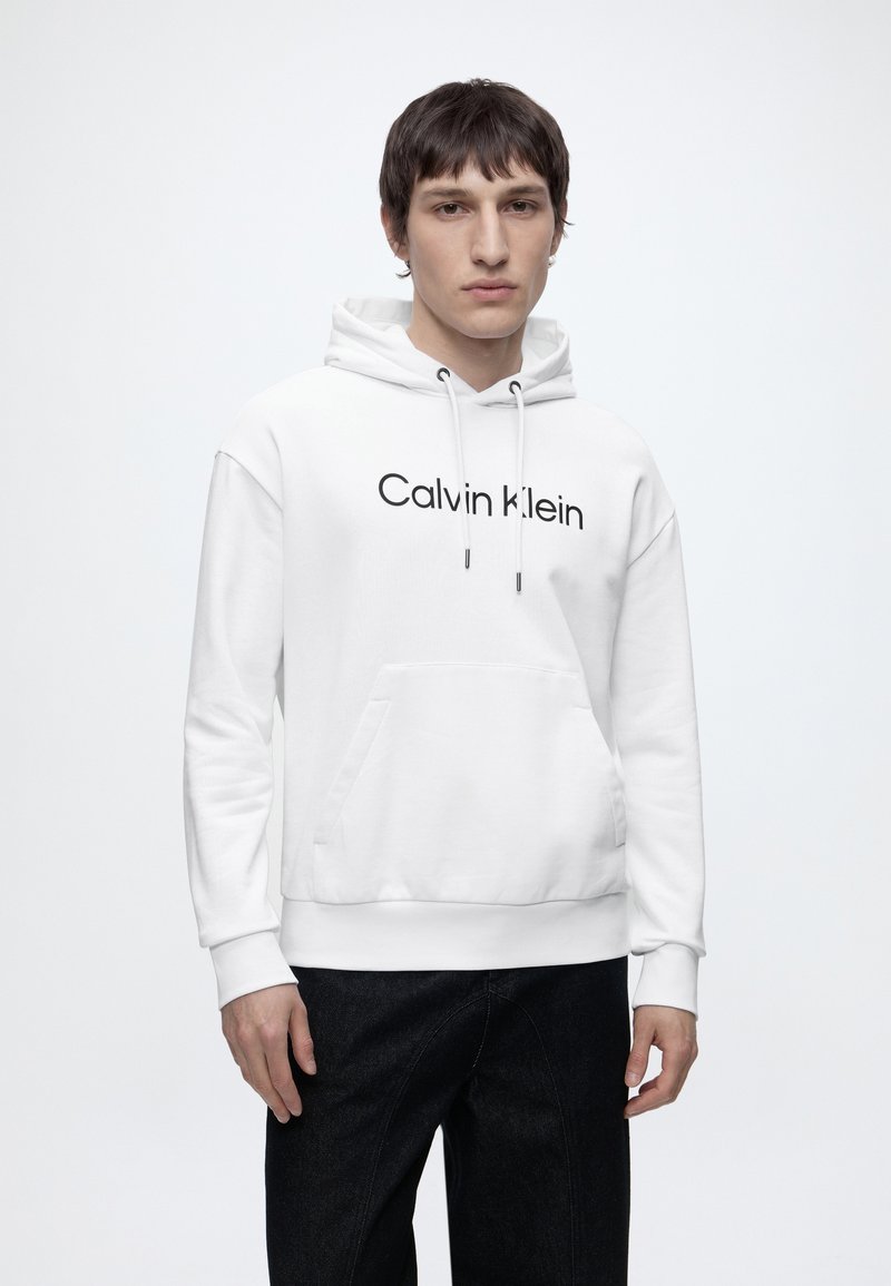Fiatal férfi, szemben, fehér Calvin Klein kapucnis pulóvert és fekete nadrágot visel, egyszínű, világos háttér előtt.