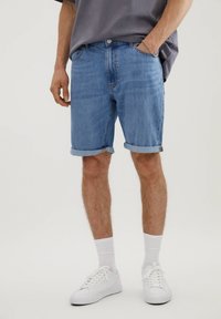 Shorts de mezclilla azul claro con puños enrollados, cinco bolsillos y un corte relajado. Combinados con zapatillas blancas y calcetines blancos acanalados.