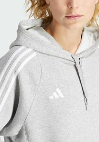Sweat à capuche gris avec une capuche prononcée, featuring des rayures blanches, une fermeture éclair à l'avant et le logo Adidas en blanc sur la poitrine.