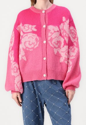 Femme portant un cardigan en tricot rose avec des motifs floraux rose clair et des boutons blancs, associé à un pantalon bleu orné de petites perles.