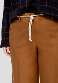 Pantalones marrones con una textura suave, que cuentan con un cinturón de cuerda blanco. La parte superior tiene una base azul marino oscura con un patrón de cuadrícula naranja.