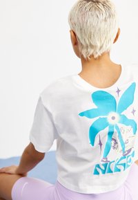 Valkoinen lyhythihainen t-paita, jossa on suuri sininen palmupuu graafinen kuva ja violetit yksityiskohdat selässä. Kangas näyttää pehmeältä ja rentouttavalta.