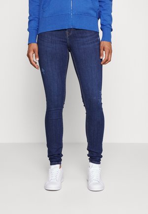 Skinny dark blue jeans with a slight fade, featuring a classic five-pocket design. Paired with white sneakers and a blue zip-up hoodie.