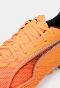 Fotbalová bota v zářivě oranžové barvě s texturovaným svrškem, výraznými černými a šedými grafickými prvky a kontrastními tkaničkami.