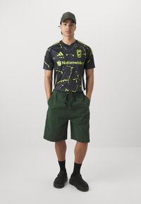 Camiseta de fútbol negra con un patrón salpicado en amarillo neón, pantalones cortos verdes, zapatos negros y una gorra verde oscura. Diseño sencillo con una estética deportiva.