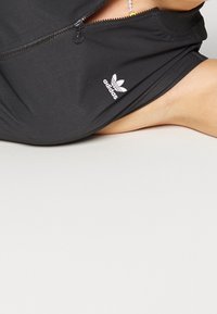 Svart zip-up plagg med slät textur, prytt med en vit Adidas-logga och en tajt passform. Stretchigt och lätt material.