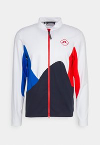 Vit, röd och blå zip-jacka med hög krage, djärva färgblock och en logotyp på bröstet. Tillverkad av lätt material.
