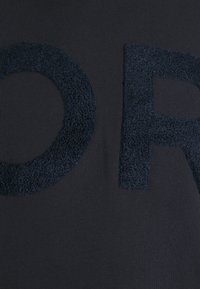 Prenda de color azul marino oscuro con letras de tela texturizada y suave que deletrean "OR". El material de fondo liso contrasta con el detalle de las letras elevadas.