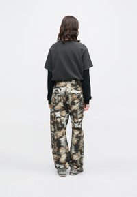 Άτομο με μεσαίου μήκους σκούρα μαλλιά φοράει επικαλυμμένα μαύρα τοπ, καφέ και γκρίζα παντελόνια με αφηρημένο γεωμετρικό σχέδιο και γκρίζα μποτάκια πεζοπορίας, γυρισμένο προς τα πίσω.