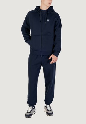 Chándal azul marino que incluye una sudadera con cremallera y cordones, y pantalones a juego. Tela suave con un corte relajado y un ligero brillo.