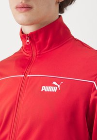 Červená športová bunda vyrobená z hladkej tkaniny s vysokým golierom, so zvodnými bielymi pruhmi a bielym logom Puma na ľavej strane.