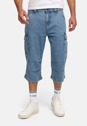 Homme portant un short cargo en jean bleu, des baskets blanches et des chaussettes blanches mi-mollet avec des rayures noires, debout devant un fond uni.